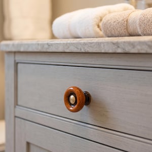 May include: A round, polished wooden drawer knob with a warm, amber-toned grain and a central metallic gold-colored circular inlay. The knob is mounted on a light gray, wood-grained cabinet drawer featuring a clean, horizontal rectangular design.
