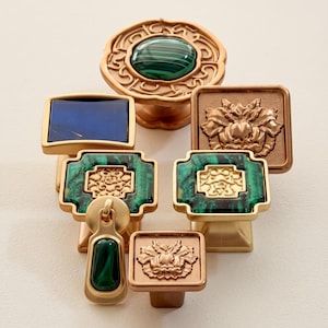 May include: A collection of six decorative cabinet knobs in various shapes and finishes. The set includes round, square, and rectangular knobs featuring gold-toned metal, green malachite-style inlays, a deep blue rectangular piece, and floral reliefs.