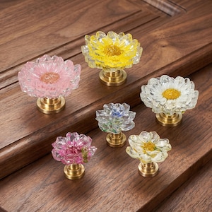 Peut inclure: Six boutons de placard en forme de fleur avec des pétales en résine transparente et des bases en métal doré. Les boutons présentent des centres floraux incrustés de différentes couleurs, dont rose, jaune, blanc et bleu, disposés sur du bois.