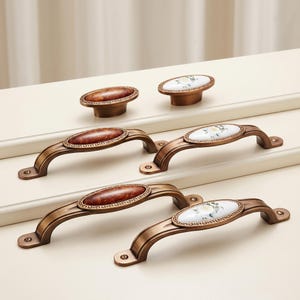 May include: A collection of six cabinet hardware pieces featuring two oval knobs and four arched handles. The hardware has a brushed bronze finish with a Greek key border. Three pieces feature a brown marble-like inlay, and three feature a white floral inlay.