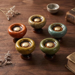 May include: Five round ceramic cabinet knobs with a glossy, speckled glaze in orange, tan, teal, yellow, and green. Each knob features a central circular brass-toned metal inlay set within a recessed dark brown center, mounted on a dark base.