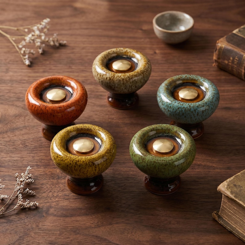 May include: Five round ceramic cabinet knobs with a glossy, speckled glaze in orange, tan, teal, yellow, and green. Each knob features a central circular brass-toned metal inlay set within a recessed dark brown center, mounted on a dark base.