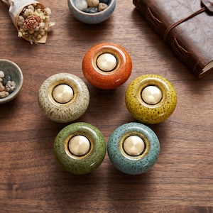 May include: Five round ceramic cabinet knobs arranged in a circle, each featuring a speckled glaze in orange, yellow, green, blue, and cream. Each knob has a smooth, polished brass-colored circular metal insert centered on its top surface.