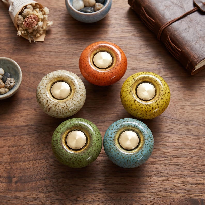 May include: Five round ceramic cabinet knobs arranged in a circle, each featuring a speckled glaze in orange, yellow, green, blue, and cream. Each knob has a smooth, polished brass-colored circular metal insert centered on its top surface.