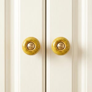 May include: Two round cabinet knobs featuring a speckled mustard yellow ceramic finish with a smooth, circular brass-toned metal center insert. The knobs are mounted on a white paneled door surface, providing a contrast in color and texture.