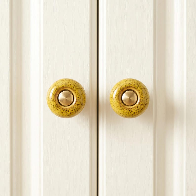 May include: Two round cabinet knobs featuring a speckled mustard yellow ceramic finish with a smooth, circular brass-toned metal center insert. The knobs are mounted on a white paneled door surface, providing a contrast in color and texture.