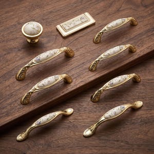 Può includere: Una collezione di ferramenta dorata per mobili composta da un pomello rotondo, una piastra rettangolare e sei maniglie ad arco per cassetti. Ogni pezzo presenta un inserto in ceramica decorato con un delicato motivo floreale di piccole foglie blu e verdi.