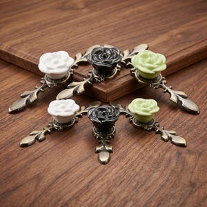 May include: A set of five drawer pulls featuring rose-shaped knobs in white, black, and light green. Each knob is mounted on an ornate, antique brass-finished metal base with a leaf-like design extending from both sides of the central rose element.