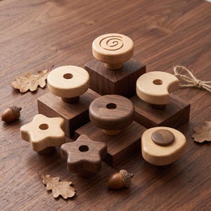 Könnte beinhalten: Eine Sammlung von Holzknöpfen in verschiedenen Formen, darunter Sterne, Kreise und ein Spiralmuster, gefertigt aus hellem und dunklem Holz. Die Knöpfe sind auf quadratischen Holzblöcken montiert, umgeben von Eichenblättern und kleinen Eicheln.