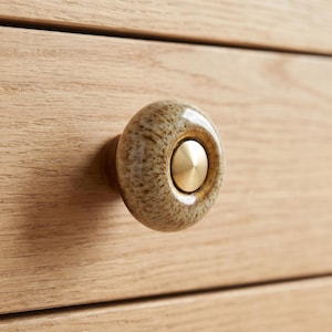 May include: A round cabinet knob featuring a speckled beige and brown ceramic body with a smooth, glossy finish. The center of the knob is inset with a circular, brushed gold-toned metal disc that has a slightly raised, conical center point.