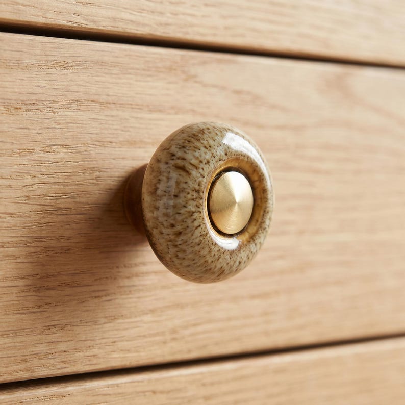 May include: A round cabinet knob featuring a speckled beige and brown ceramic body with a smooth, glossy finish. The center of the knob is inset with a circular, brushed gold-toned metal disc that has a slightly raised, conical center point.