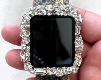 May include: A silver Apple Watch with a clear face and a diamond-encrusted bezel. The watch is on a brown and beige patterned band with a bee design.