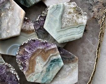 May include: A collection of hexagonal agate coasters with various patterns of white, green, purple, and brown. The coasters are arranged on a larger, light pink agate coaster with a gold trim.