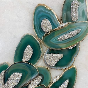 May include: A collection of green agate slices with natural banding, each featuring a gold-toned metallic edge and a cluster of clear, sparkling crystals embedded on the surface. The stones vary in shape and size, displaying deep emerald hues.