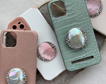 May include: Four different phone cases with a variety of colors and textures. Each case has a decorative ring holder with a pearl-like center and rhinestones around the edge. The cases are arranged on a light gray surface.