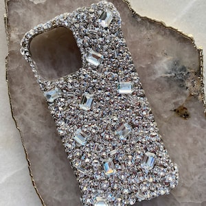 May include: A clear phone case covered in small and large rhinestones. The case is designed for a smartphone with a camera bump.