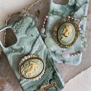 May include: Two phone cases with green marble patterns. One case features a gold-rimmed cameo of a mermaid surrounded by small white pearls and a gold plaque reading IDEAL OF SWEDEN. The other case has a gold-framed mermaid cameo and a beaded bracelet.