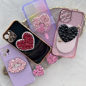 May include: Five different phone cases with jeweled heart and lip designs. The cases are in various colors, including black, pink, purple, and light blue. The jeweled designs are made of rhinestones and glitter.