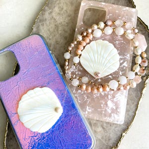May include: Two iridescent phone cases with a white seashell design. One case has a pink and white beaded strap with a pearl accent.