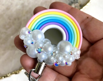 Puede incluir: Un llavero con forma de arcoíris con una nube blanca hecha de perlas y pedrería. El arcoíris está hecho de múltiples capas de colores pastel, incluyendo rosa, amarillo, verde, azul y morado. El llavero tiene un clip giratorio plateado y un llavero plateado.