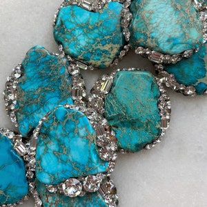 Può includere: Primo piano di una collana con grandi pietre turchesi irregolari. Ogni pietra è bordata da un bordo di cristalli chiari e scintillanti. Le pietre turchesi hanno un aspetto marmorizzato con sfumature di blu e marrone.