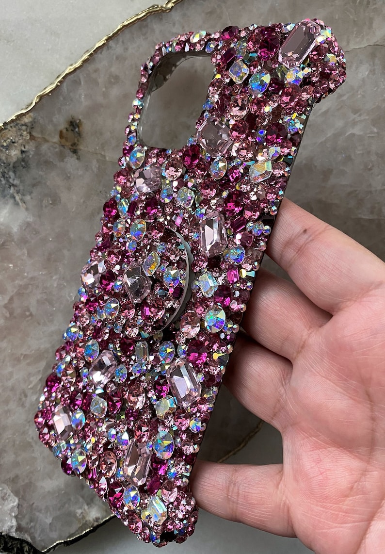 May include: A pink and iridescent phone case covered in rhinestones with a clear phone grip.