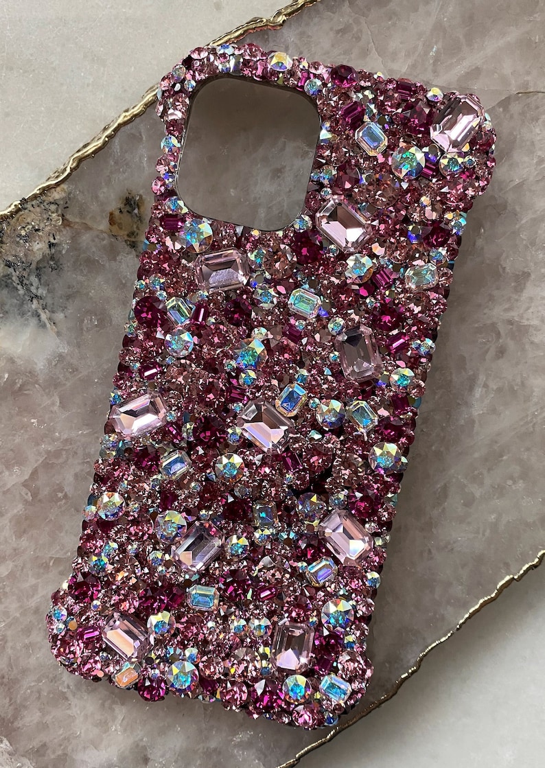 May include: A pink and iridescent phone case covered in rhinestones. The case is designed to fit a smartphone with a camera cutout.
