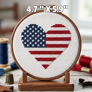 May include: A circular wooden embroidery hoop containing white Aida fabric with a cross-stitch heart design featuring the American flag pattern of red and white stripes and a blue field with white stars. The text 4.7" X 5.9" is visible.