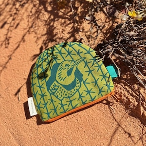 May include: A small, green and teal patterned pouch with an orange trim and a teal zipper pull. The pouch features a teal bird design. The pouch is lying on a bed of red sand with yellow flowers and dry brush in the background.