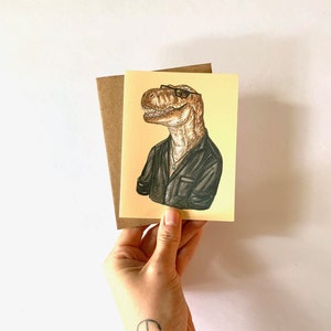 May include: Greeting card featuring a Tyrannosaurus Rex wearing glasses and a black shirt. The dinosaur illustration is on a yellow background, with a brown envelope behind it. The card is held in a hand with an anchor tattoo.