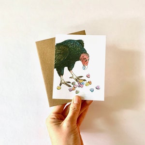 May include: A greeting card featuring a black and red vulture with a pink candy heart in its beak. The vulture is surrounded by a scattering of colorful candy hearts.