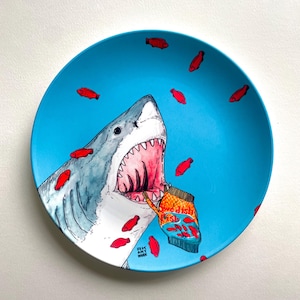 May include: A round, blue ceramic plate featuring a shark with an open mouth, holding a bag of Swedish Fish candy. Red candy fish are scattered around the shark. The plate has the text "RK ART WORK".
