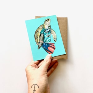 May include: A greeting card featuring a cartoon turtle wearing a diving mask, snorkel, and flippers. The turtle is on a turquoise background. The card is held with a brown envelope.