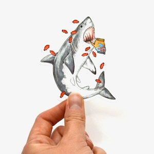 May include: A white shark with an open mouth holding a box of orange goldfish crackers. The shark is surrounded by orange goldfish crackers.