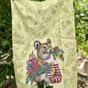 May include: A yellow tea towel with a floral pattern and a cartoon animal holding a bouquet of flowers. The animal is wearing a red and white striped sweater.