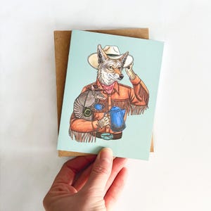 May include: A greeting card featuring a coyote wearing a cowboy hat and a brown vest, holding a blue coffee pot. A small armadillo is sitting on the coyote's shoulder.
