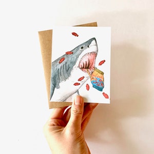 May include: A white shark with an open mouth is depicted in a watercolor illustration. The shark is eating a bag of goldfish crackers, with red goldfish crackers scattered around it.