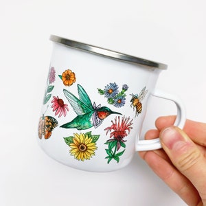 May include: White enamel mug with a silver rim and a white handle. The mug is decorated with colorful illustrations of a hummingbird, butterfly, bee, and various flowers. The mug is held by a hand.