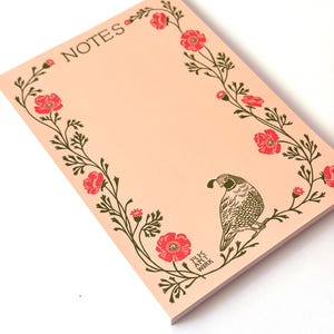 May include: A rectangular notepad with a light peach background featuring the word NOTES printed in dark green. The border consists of dark green vines with red poppy flowers and a single quail illustration. The text RK ART WORK is visible.