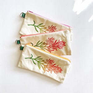 Op de afbeelding: Drie canvas etuis met bloemmotieven. Elke etui heeft een rits in een andere kleur: roze, oranje en geel. Het bloemmotief is een verloop van groen, oranje en rood. Elke etui heeft een groene lus.