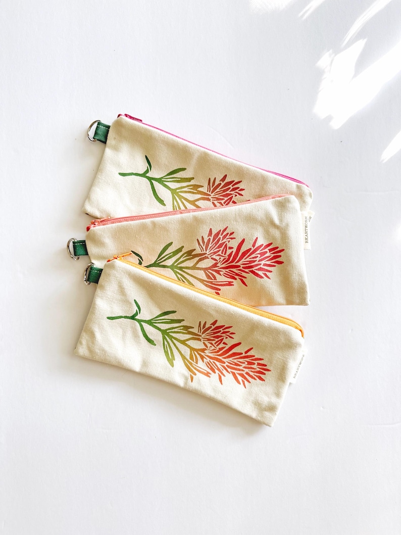 Op de afbeelding: Drie canvas etuis met bloemmotieven. Elke etui heeft een rits in een andere kleur: roze, oranje en geel. Het bloemmotief is een verloop van groen, oranje en rood. Elke etui heeft een groene lus.