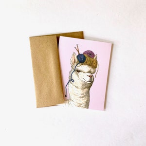 May include: A pink card with a white alpaca wearing a hat made of yarn balls and knitting needles. The alpaca is looking to the right.