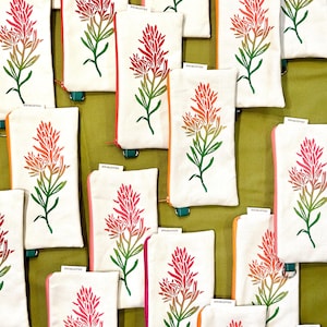 Op de afbeelding: Witte canvas pouches met een kleurrijk bloemmotief. De pouches hebben een rode, roze en oranje bloem met groene bladeren en stelen. De ritsen en randen zijn in verschillende kleuren, waaronder roze, oranje en groen.