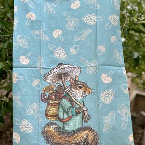 May include: A blue kitchen towel with a white mushroom pattern. A squirrel wearing a blue sweater and carrying a basket of mushrooms is walking under a mushroom umbrella.