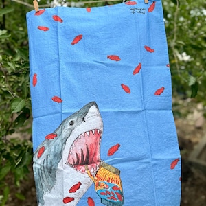 May include: A blue tea towel with a white great white shark with an open mouth eating a bag of Swedish Fish. The bag of Swedish Fish is red and white with the words "Swedish Fish" printed on it. The background of the towel is light blue with red fish printed on it.
