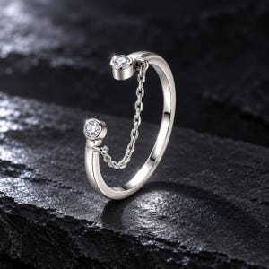 Pode incluir: Um anel aberto de tom prateado com duas extremidades circulares, cada uma cravejada com uma pedra preciosa redonda e transparente. Uma delicada corrente prateada conecta as duas extremidades do anel, criando uma ponte sobre a abertura.