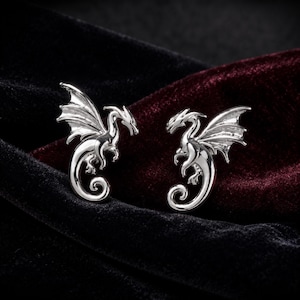 Può includere: Un paio di orecchini in metallo color argento a forma di drago. Ogni orecchino raffigura un drago dettagliato con ali spiegate, coda ricurva e testa appuntita, realizzato con una finitura metallica lucida e riflettente dai contorni nitidi.