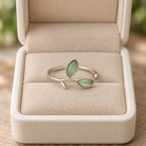 May include: A silver-toned ring featuring two smooth, light green oval-shaped stones arranged to resemble leaves on a branch. The ring is displayed inside a beige, rectangular jewellery box with a soft, textured interior lining and a hinged lid.