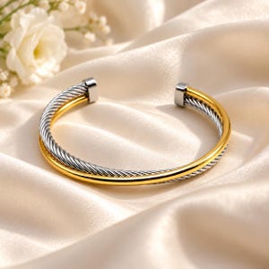 May include: A two-tone cuff bracelet featuring a polished gold-coloured band intertwined with a silver-coloured twisted cable wire band. The bracelet has a C-shaped open design with smooth, flat silver-coloured end caps at each terminal point.
