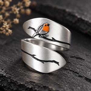 May include: A silver-toned bypass ring featuring a matte finish. The upper band displays a small bird with a bright orange chest perched on a black etched branch. The lower band continues the black etched branch design across the silver surface.